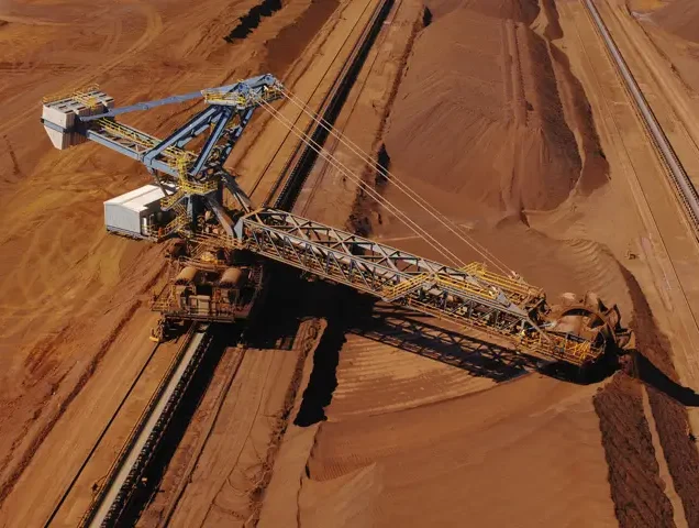 An aerial view of a large bucket-wheel excavator moving reddish-brown earth in a mining site, with piles of soil and conveyor belts visible.
