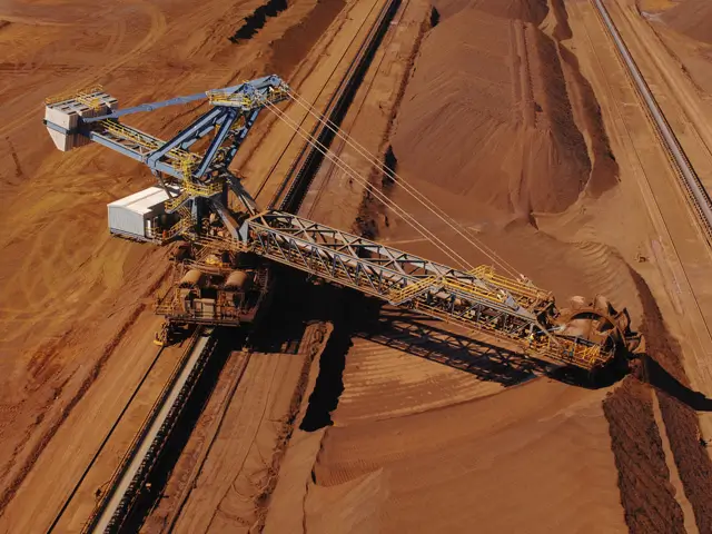 An aerial view of a large bucket-wheel excavator moving reddish-brown earth in a mining site, with piles of soil and conveyor belts visible.