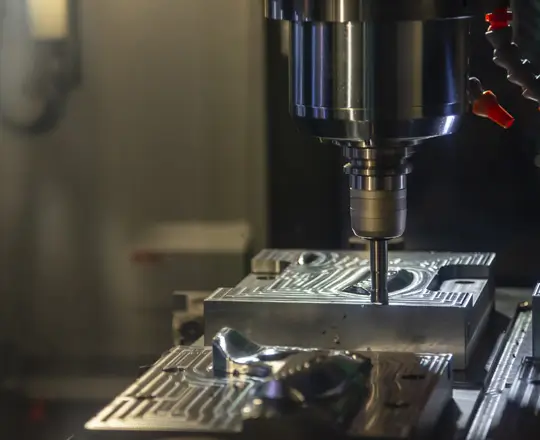A CNC machine uses a rotating drill bit to precisely carve patterns into a metal block, with intricate grooves visible on the workpiece under focused lighting.
