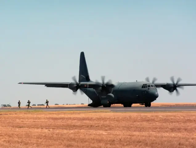 A military transport plane with spinning propellers is on a runway, while a group of soldiers walk away in the background across a dry, grassy field under a clear sky.