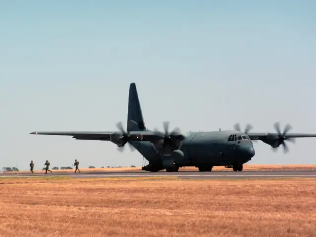 A military transport plane with spinning propellers is on a runway, while a group of soldiers walk away in the background across a dry, grassy field under a clear sky.