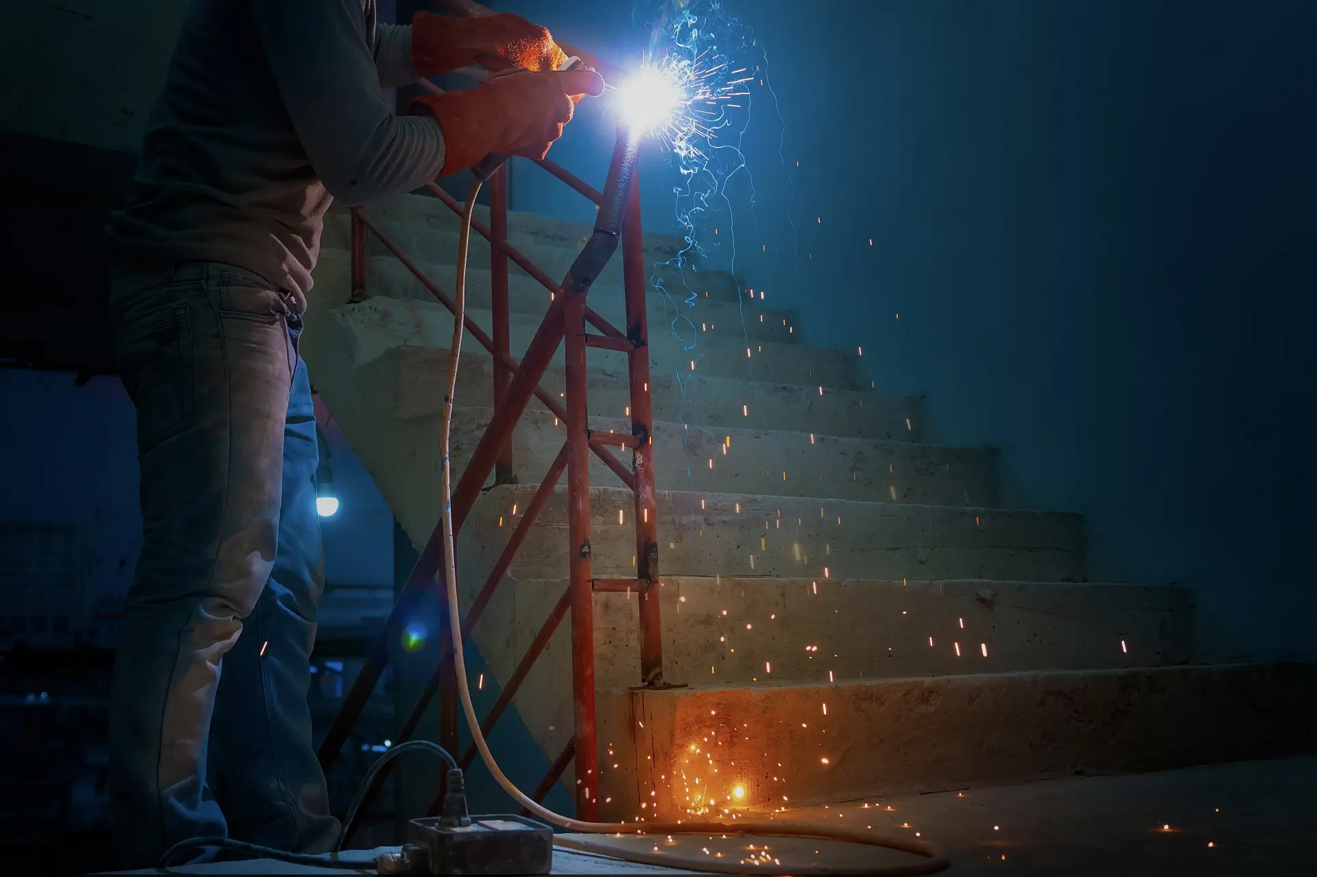 A person welding metal near a staircase, with sparks and bright light from the welding process illuminating the dimly lit area.