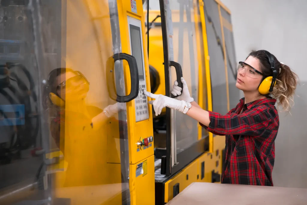 A woman wearing safety goggles, gloves, and ear protection operates the control panel of a yellow industrial machine in a factory setting.