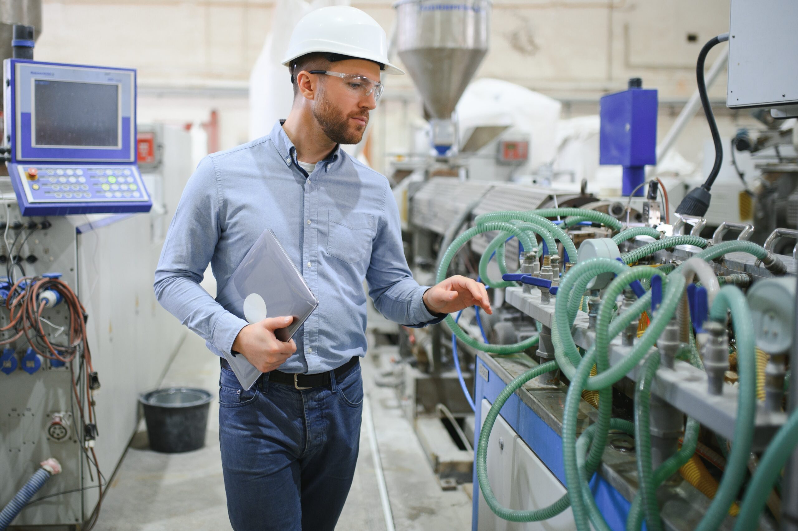 Man in safety gear inspects industrial machinery while holding a tablet in a factory setting. Various cables, tubes, and control panels are visible around him.